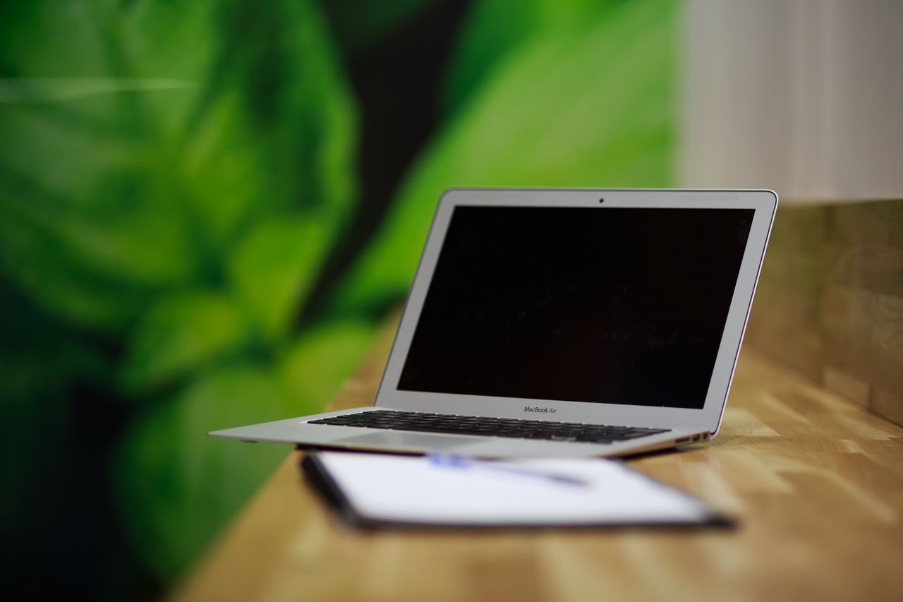 Offerings A sleek silver laptop sits open on a wooden desk with a green backdrop, illustrating a modern workspace.